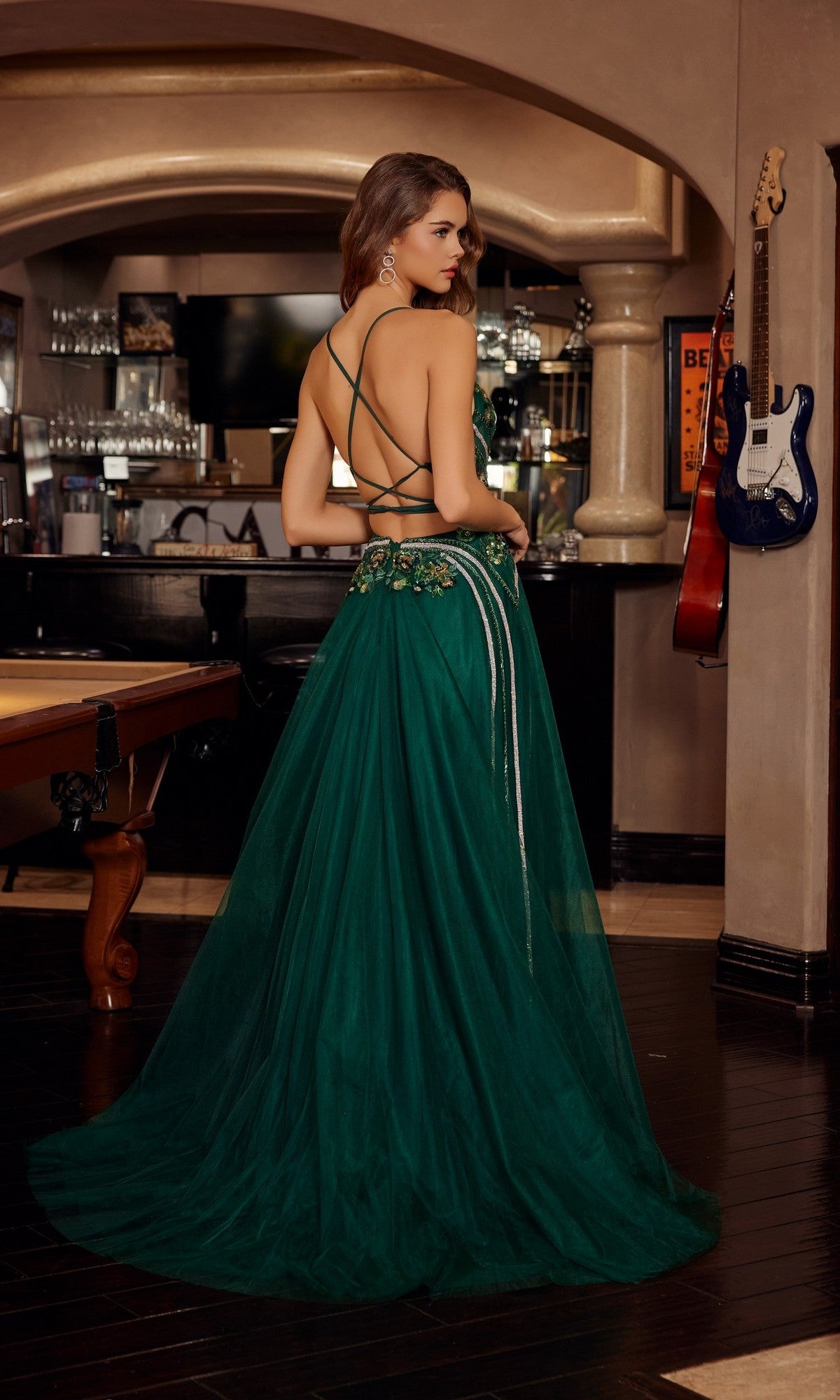 A woman models the Nox Anabel G1795 long prom dress, featuring an elegant green open-back design, while standing in a bar area with a pool table and guitars displayed on the wall.